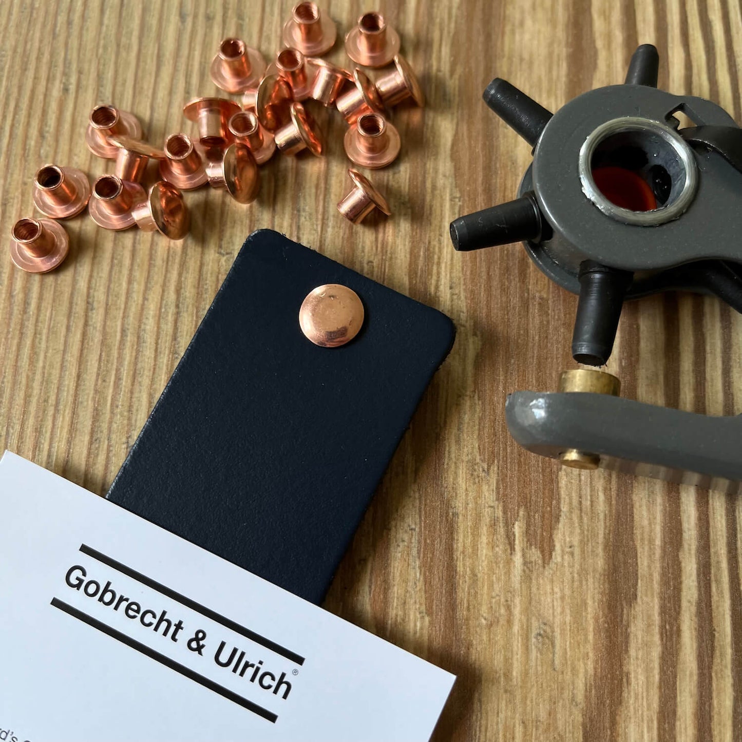 Copper binding screws scattered on a wooden surface, a silver binding screw fastened to a piece of dark blue leather, and a leather hole punch