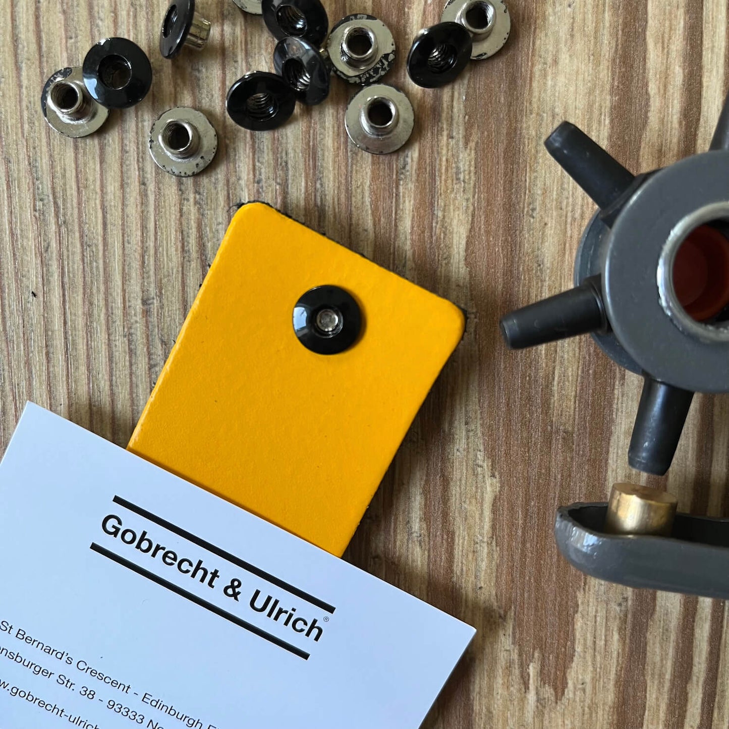 Short black binding screws scattered on a wooden surface, a silver binding screw fastened to a piece of bright yellow leather, and a leather hole punch