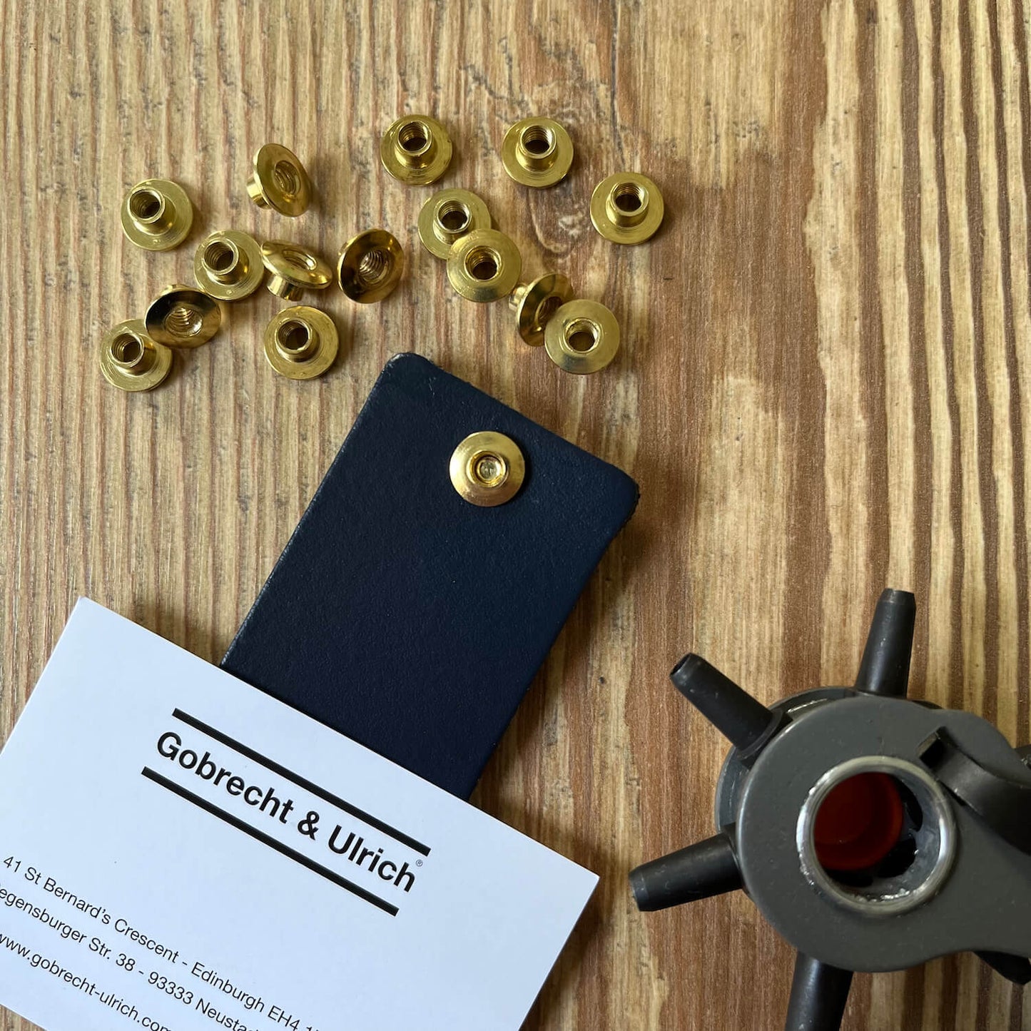 Short brass binding screws scattered on a wooden surface, a silver binding screw fastened to a piece of dark blue leather, and a leather hole punch