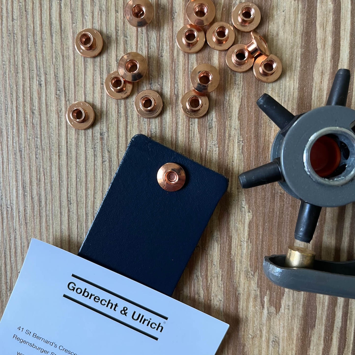 Short copper binding screws scattered on a wooden surface, a silver binding screw fastened to a piece of dark blue leather, and a leather hole punch