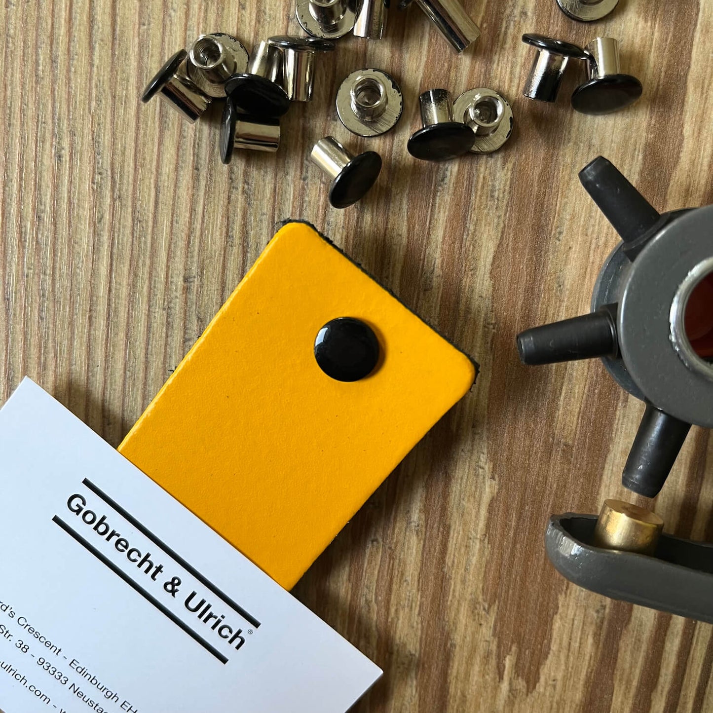 Black binding screws scattered on a wooden surface, a silver binding screw fastened to a piece of bright yellow leather, and a leather hole punch