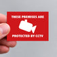 A hand holding a red rectangular vinyl sticker that reads 'THESE PREMISES ARE PROTECTED BY CCTV' in white text with a white CCTV camera icon in the center, against a plain light gray background.