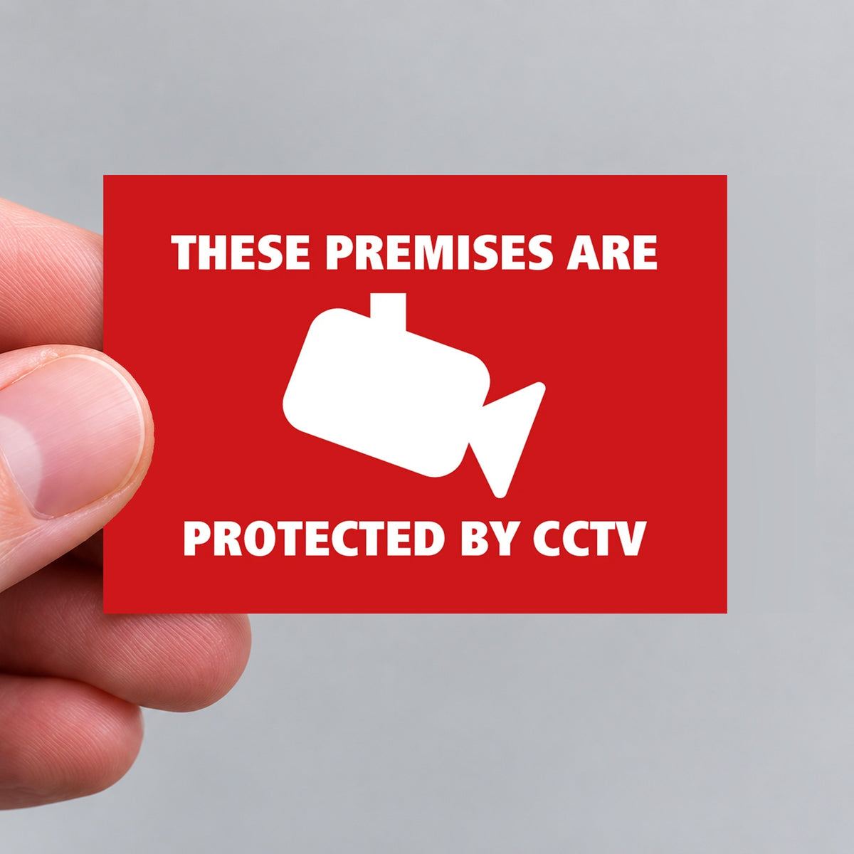 A hand holding a red rectangular vinyl sticker that reads 'THESE PREMISES ARE PROTECTED BY CCTV' in white text with a white CCTV camera icon in the center, against a plain light gray background.
