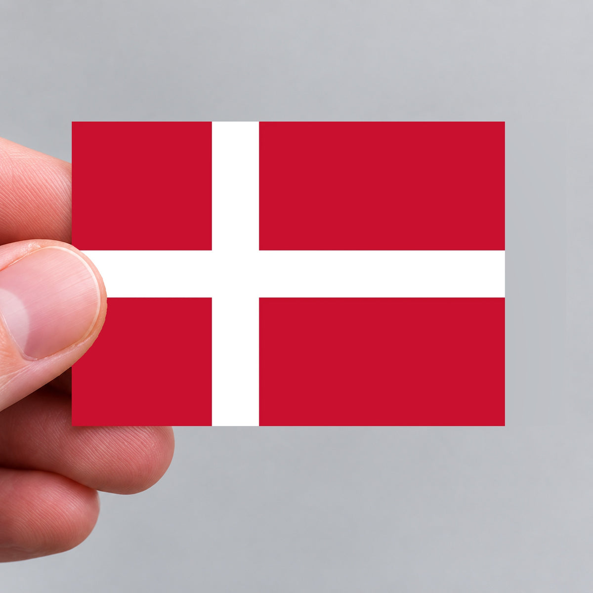 A small rectangular sticker featuring the Danish flag, held between fingers against a plain light gray background.