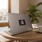 Laptop with a jolly roger sticker on a wooden table with coffee cups and a plant.