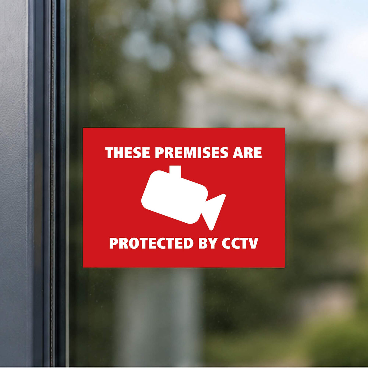 A red rectangular vinyl sticker with white text reading 'THESE PREMISES ARE PROTECTED BY CCTV' and a white CCTV camera icon, shown attached to a glass door with an outdoor blurred background.